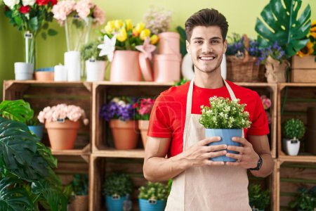 Foto de Joven hombre hispano trabajando en floristería sosteniendo planta con aspecto positivo y feliz de pie y sonriendo con una sonrisa confiada mostrando los dientes - Imagen libre de derechos