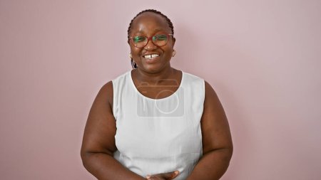 Femme afro-américaine confiante et souriante portant des lunettes, ayant une ambiance amusante et positive, se tenant décontractée sur un fond rose isolé.