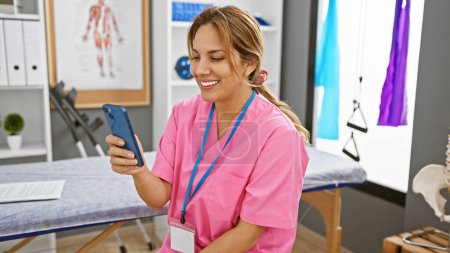 Photo for A cheerful latina healthcare worker in pink scrubs using a smartphone in a modern rehabilitation clinic. - Royalty Free Image