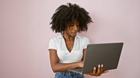 Téléchargez les photos : Femme afro-américaine utilisant un ordinateur portable avec une expression sérieuse sur fond rose isolé - en image libre de droit