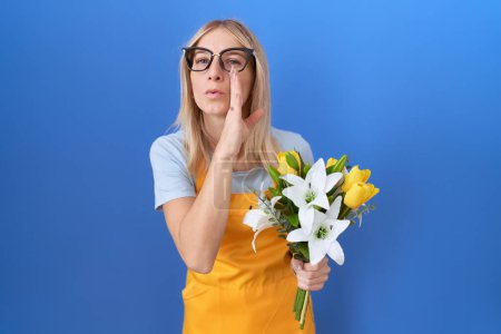Photo for Young caucasian woman wearing florist apron holding flowers hand on mouth telling secret rumor, whispering malicious talk conversation - Royalty Free Image