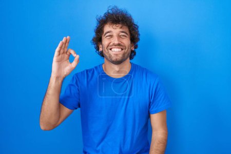 Foto de Hispanic young man standing over blue background smiling positive doing ok sign with hand and fingers. successful expression. - Imagen libre de derechos
