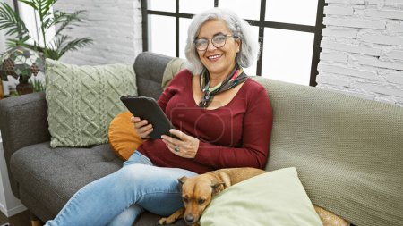 Téléchargez les photos : Femme mûre souriante aux cheveux gris se relaxant sur un canapé avec son chien, tenant une tablette dans un salon confortable. - en image libre de droit