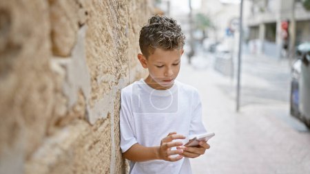 Photo for Adorable blond boy, engrossed in a serious conversation on smartphone, standing outdoors on city street. cute child artistically leaning against wall, fully concentrated on texting. - Royalty Free Image