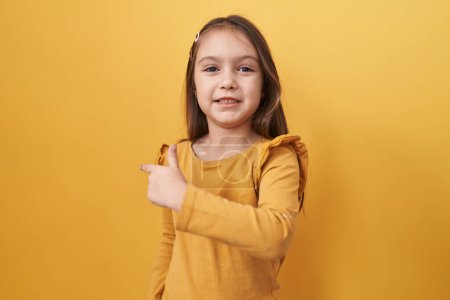 Photo for Adorable hispanic girl confidently standing, cheerfully pointing up to the side with her hand and finger against a yellow isolated background, radiating with a beautiful natural smile - Royalty Free Image