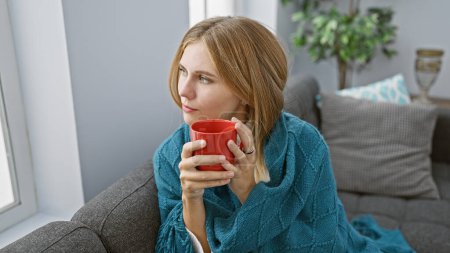 Téléchargez les photos : Femme blonde aux yeux bleus dégustant du café à la maison, enveloppée dans une couverture sarcelle sur un canapé gris. - en image libre de droit