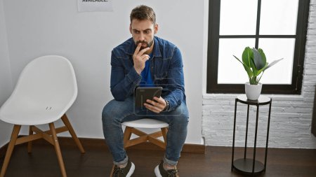 Photo for Young hispanic adult with a beard, deep in thought, sitting relaxed in the waiting room, using his touchpad device amidst the interior lobby background - Royalty Free Image