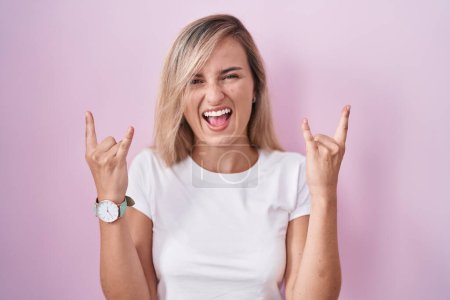 Photo for Young blonde woman standing over pink background shouting with crazy expression doing rock symbol with hands up. music star. heavy music concept. - Royalty Free Image