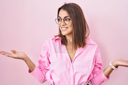 Téléchargez les photos : Jeune femme hispanique portant des lunettes debout sur fond rose souriant montrant les paumes ouvertes des deux mains, présentant et comparant la publicité et l'équilibre - en image libre de droit