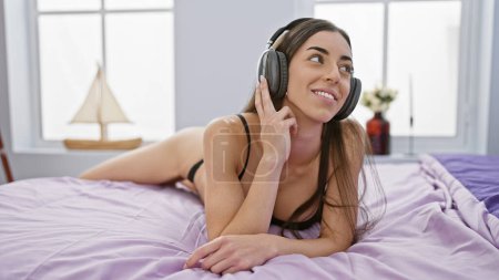 Photo for Beautiful young hispanic woman awakes in the morning, lies in bed, sensually enjoying music in her comfortable room, oozing confidence with her relaxed body language and sexy lingerie. - Royalty Free Image