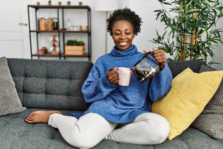 Téléchargez les photos : African american woman pouring coffee on cup sitting on sofa at home - en image libre de droit