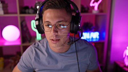 Photo for Handsome hispanic man wearing headphones in a vibrant gaming room with neon lights at night - Royalty Free Image