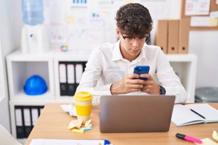 Téléchargez les photos : Jeune adolescent hispanique travailleur d'entreprise utilisant un ordinateur portable et un smartphone au bureau - en image libre de droit