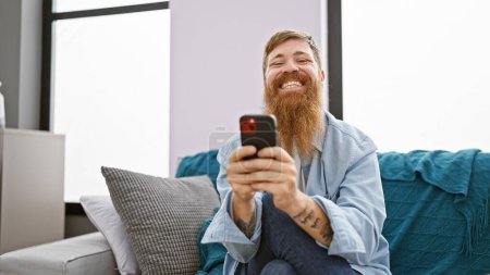 Photo for Handsome young redhead man cheerfully using his smartphone while sitting on the sofa in the living room of his home, spreading joy and positive vibes through texting and the online world - Royalty Free Image