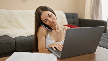 Photo for Joyous young hispanic woman, beautifully smiling, basks in comfort as she enjoys engaging talk via smartphone and laptop on the floor at home - Royalty Free Image