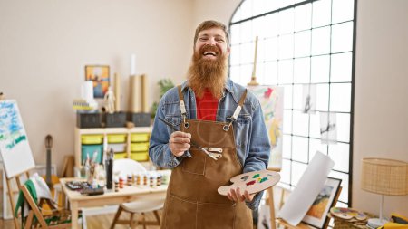 Téléchargez les photos : Jeune homme rousse confiant artiste pinceau à la main, souriant joyeusement dans un studio d'art d'intérieur - en image libre de droit