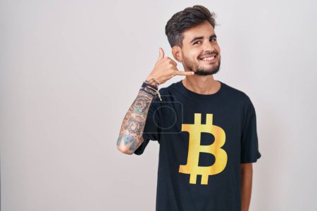 Foto de Joven hombre hispano con tatuajes con camiseta bitcoin sonriendo haciendo gesto telefónico con la mano y los dedos como hablar por teléfono. comunicar conceptos. - Imagen libre de derechos