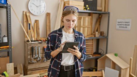 Téléchargez les photos : Une jeune femme concentrée utilise une tablette dans un atelier bien équipé avec des outils de travail du bois et des panneaux de sécurité. - en image libre de droit