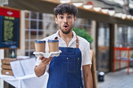 Foto de Hombre árabe con barba llevando delantal de camarero en la terraza del restaurante asustado y sorprendido con la boca abierta para sorpresa, cara de incredulidad - Imagen libre de derechos