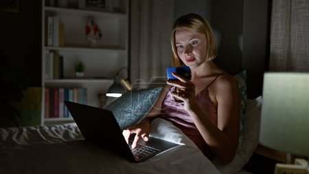 Téléchargez les photos : Jeune femme blonde faisant du shopping avec ordinateur portable et carte de crédit assise sur le lit dans la chambre - en image libre de droit