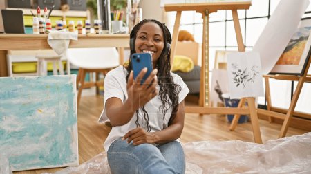Téléchargez les photos : Une femme afro-américaine prend un selfie avec son smartphone, souriant avec des écouteurs, dans un studio d'art lumineux et créatif. - en image libre de droit