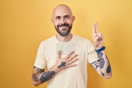 Téléchargez les photos : Homme hispanique avec des tatouages debout sur fond jaune souriant jurant avec la main sur la poitrine et les doigts vers le haut, faisant un serment de fidélité - en image libre de droit
