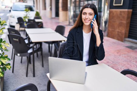 Photo for Young beautiful hispanic woman talking on smartphone using laptop sitting on table at coffee shop terrace - Royalty Free Image