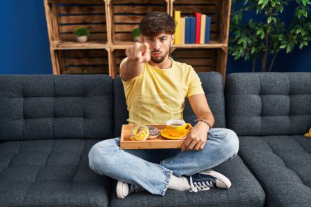 Foto de Hombre árabe con barba sentado en el sofá en casa desayunando apuntando con el dedo a la cámara y a ti, gesto confiado y serio - Imagen libre de derechos