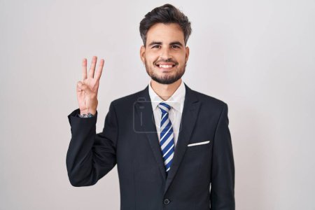 Téléchargez les photos : Jeune homme hispanique avec des tatouages portant un costume d'affaires et une cravate montrant et pointant vers le haut avec les doigts numéro trois tout en souriant confiant et heureux. - en image libre de droit