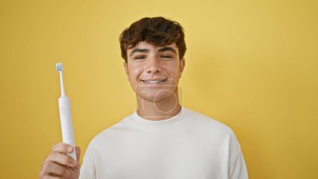 Foto de Joven adolescente hispano confiado, sonriendo ampliamente con un cepillo de dientes eléctrico sobre un fondo aislado amarillo vibrante - Imagen libre de derechos