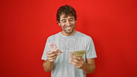Foto de Joven radiante, contando con confianza su riqueza en billetes argentinos, aislado sobre un vibrante fondo rojo - Imagen libre de derechos