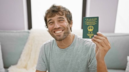 Photo for Cheerful young man, casually sitting on the living room sofa, joyfully holds his macao passport indoor, displaying a confidence-filled, happy smile at home. - Royalty Free Image