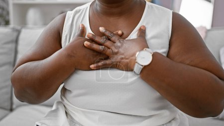 Foto de Sincera gratitud, mujer afroamericana con sobrepeso, manos en el pecho, sintiéndose agradecida en su sala de estar - Imagen libre de derechos