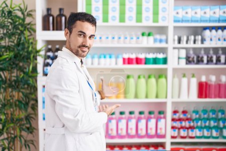 Téléchargez les photos : Bel homme hispanique travaillant à la pharmacie pharmacie invitant à entrer souriant naturel avec la main ouverte - en image libre de droit