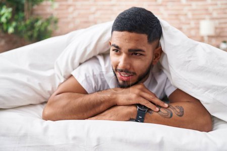 Téléchargez les photos : Jeune homme latin couché sur le lit couvrant avec drap de lit dans la chambre - en image libre de droit