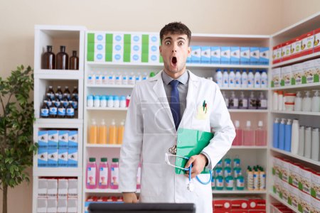 Foto de Joven hombre hispano trabajando en farmacia sosteniendo estetoscopio asustado y sorprendido con la boca abierta para sorpresa, cara de incredulidad - Imagen libre de derechos