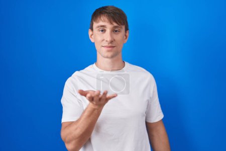 Photo for Caucasian blond man standing over blue background smiling cheerful offering palm hand giving assistance and acceptance. - Royalty Free Image