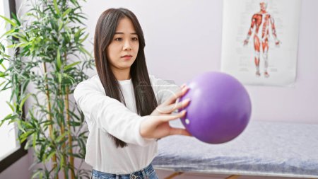 Téléchargez les photos : Une jeune femme asiatique fait des exercices avec une balle violette dans la salle de désintox d'une clinique, illustrant les soins de santé et de remise en forme. - en image libre de droit