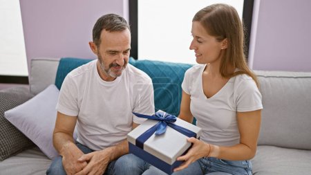 Foto de Sorpresa alegre de padre e hija, desvelando un regalo de cumpleaños juntos en el sofá de la sala de estar en casa - Imagen libre de derechos