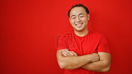 Foto de Riéndose guapo joven asiático hombre exuda confianza, de pie con brazos cruzados sobre rojo fondo, encarnando alegría y positividad - Imagen libre de derechos