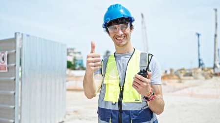 Foto de Joven arquitecto hispano sosteniendo walkie-talkie haciendo gesto de pulgar hacia arriba en el lugar de construcción - Imagen libre de derechos