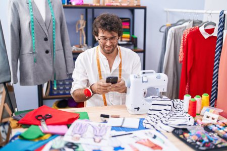 Téléchargez les photos : Jeune homme hispanique tailleur souriant confiant en utilisant smartphone à l'usine de vêtements - en image libre de droit
