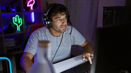 Téléchargez les photos : Un homme concentré avec des écouteurs utilise un ordinateur portable dans une salle de jeux éclairée au néon la nuit. - en image libre de droit