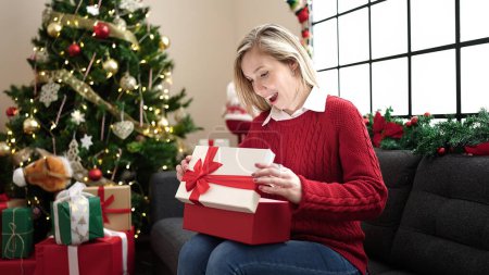 Photo for Young blonde woman unpacking gift sitting on sofa by christmas tree at home - Royalty Free Image