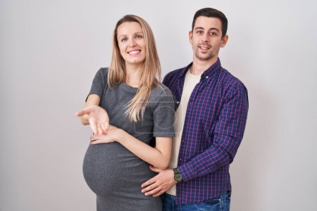 Photo for Young couple expecting a baby standing over white background smiling cheerful offering palm hand giving assistance and acceptance. - Royalty Free Image