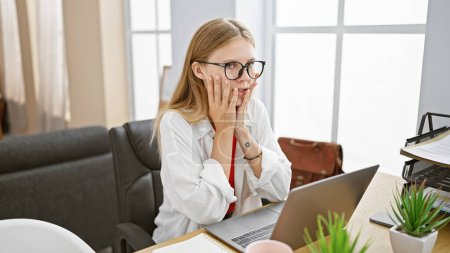 Foto de Una mujer rubia sorprendida con gafas reacciona en su escritorio de la oficina con un ordenador portátil y plantas en una habitación llena de luz natural. - Imagen libre de derechos