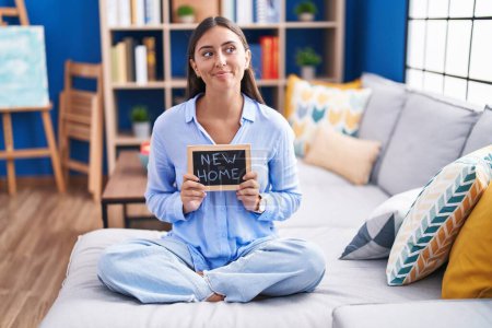 Photo for Young hispanic woman holding blackboard with new home text sitting on the sofa smiling looking to the side and staring away thinking. - Royalty Free Image
