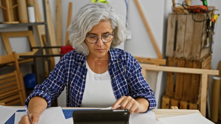 Photo for A mature woman in a workshop uses a tablet while engaged in a creative carpentry project indoors. - Royalty Free Image