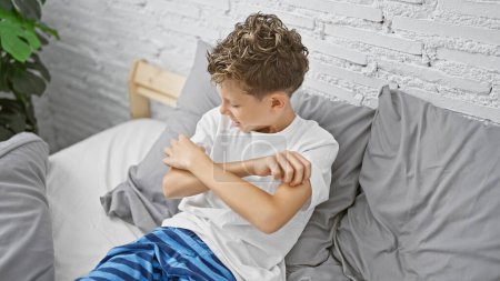 Téléchargez les photos : Adorable garçon blond assis dans la chambre à coucher, éraflure du bras qui gratte - problème d'allergie intérieure ou eczéma ? santé de l'enfant en jeu dans l'environnement familial! - en image libre de droit