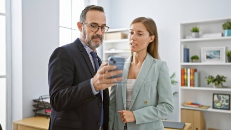 Foto de Dos trabajadores de negocios serios de pie juntos en el interior, inmersos en una conversación de trabajo en línea exitosa en el teléfono inteligente en la oficina profesional - Imagen libre de derechos